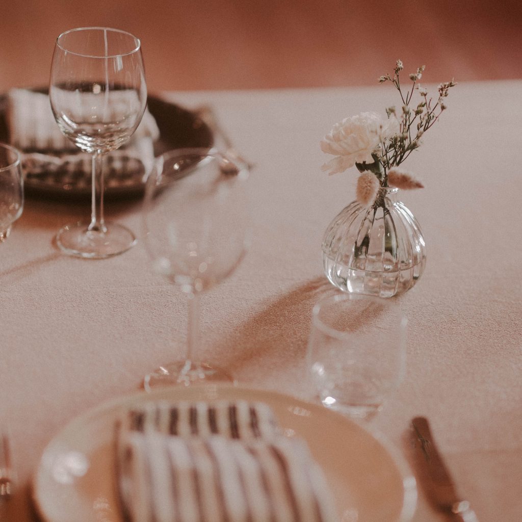 Juhlakattau pöydällä, viinilasti, lautset, aterimet ja pieni kukkakimppu.