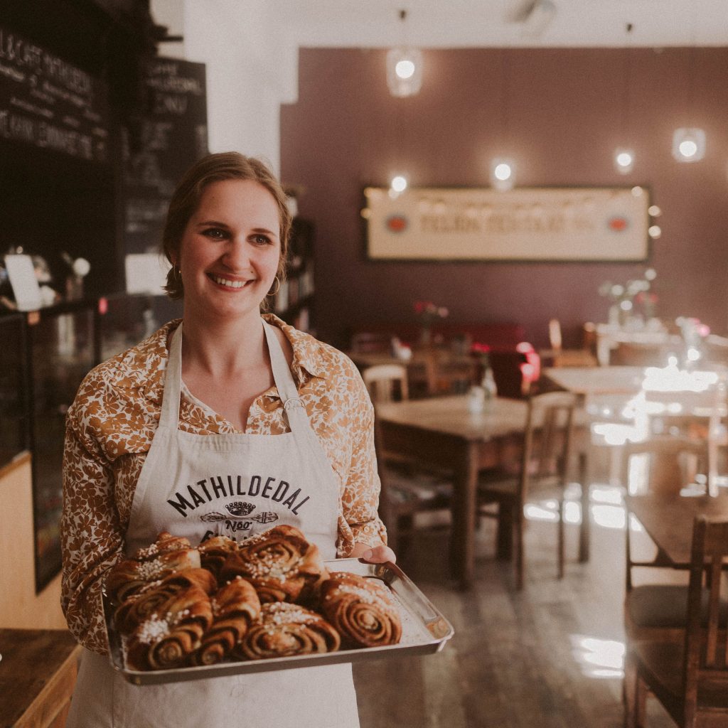 Café Mathildedalin hymyilevä tarjoilia, jolla keltakuvioinen paita ja beige esiliina ja korvapuustitarjotin kädessä.