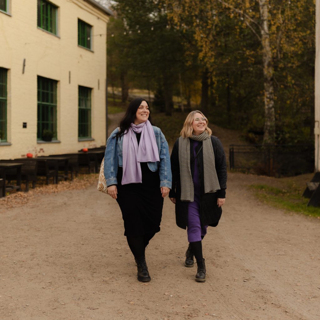 Kaksi naista kävelee kameraa kohti hiekkatietä pitkin Mathildedalin ruukkialueella. Taustalla näkyy keltainen vanha tehdasrakennus.