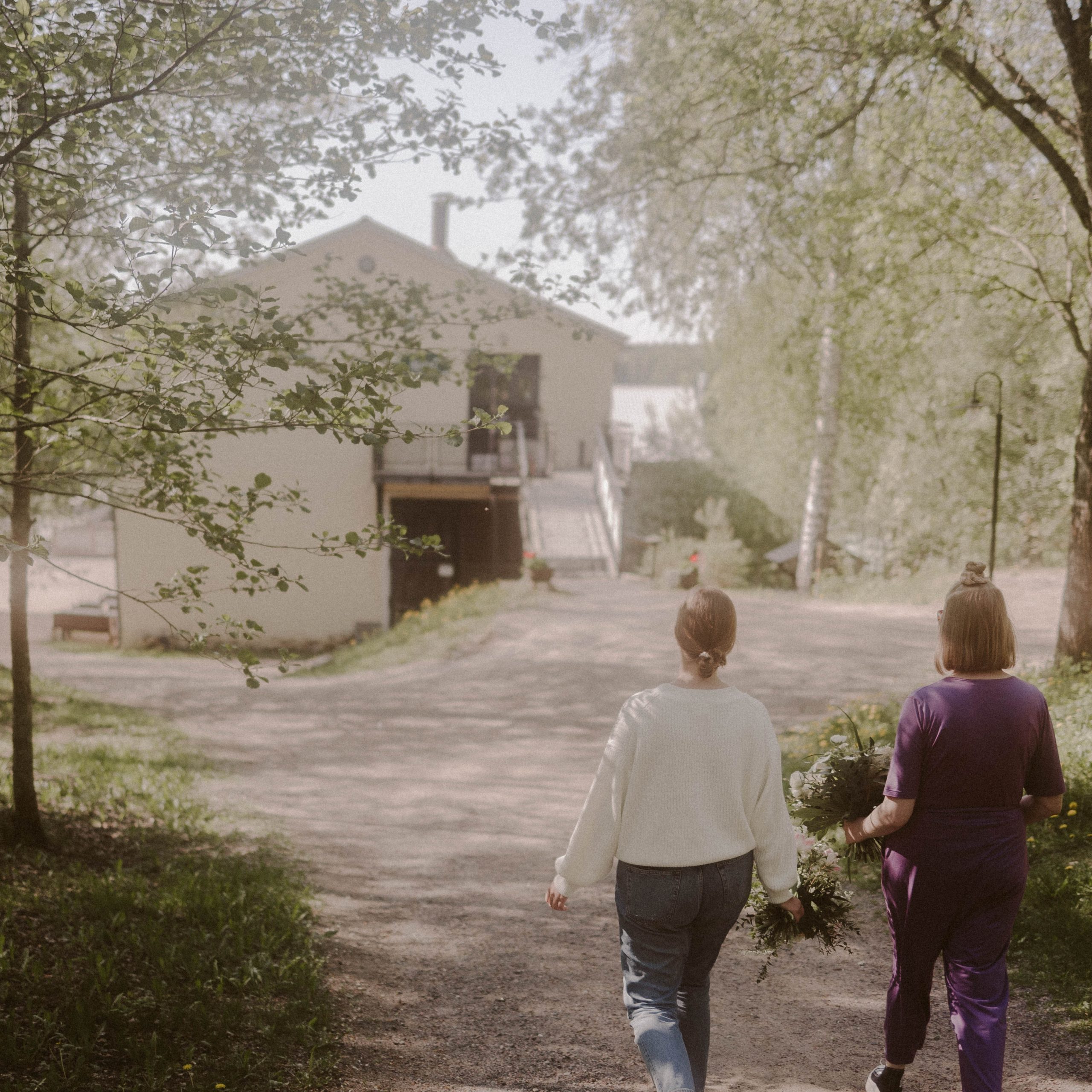 Kaksi nuorta naista kävelemällä hiekkatietä pitkin Mathildedalin Ruukkialuella. Taustalla on keltainen vanha tehdasrakennus.