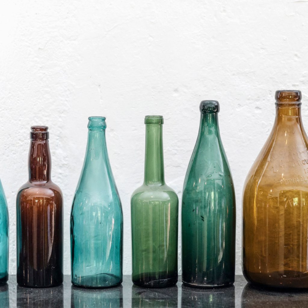 Vanhoja sinisiä, vihreitä ja ruskeita pulloja rivissä valkoisen kiviseinän edessä.