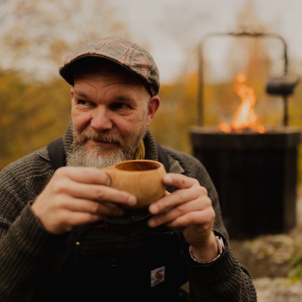 Lakkipäinen mies istuu kuksa kädessä metsässä. Taustalla on nuotio.