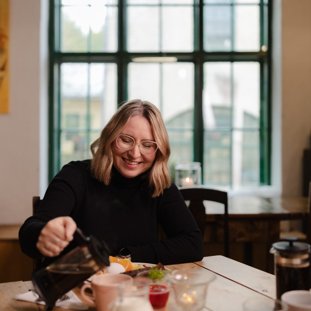 Nainen, jolla vaaleat hiukset ja silmälasit, istuu aamiaisella Café Mathildedalissa. Taustalla on iso vihreäpokainen ikkuna.