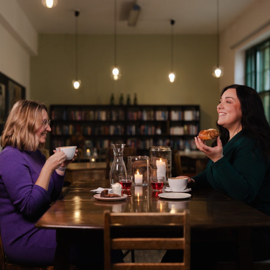 Kaksi naista istuvat vastakkain pöydän ympärillä pitämässä kokousta ja kahvittelemassa.